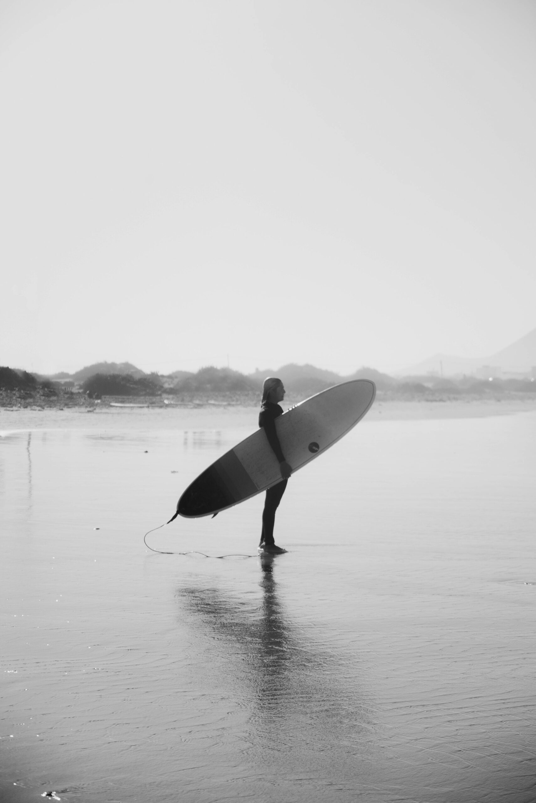 Black and white photo of a surfer with surfboard standing by calm sea at dusk.